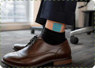 A close-up of a man's lower leg in an office, wearing navy suit trousers, black socks and brown leather derby shoes, with a teal MagicPatch grid sticker on his ankle just above the sock.