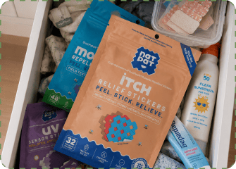 An open dresser drawer holding a MagicPatch Itch Relief Stickers pack in front of BuzzPatch and UV Sensor sticker packs, next to a container of bandages, a tube of nappy rash ointment and a bottle of kids' clear SPF50 sunscreen.