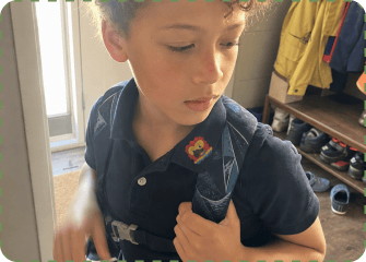 A child adjusting a backpack strap while wearing a Seasonal patch on their shirt collar in a home entryway.