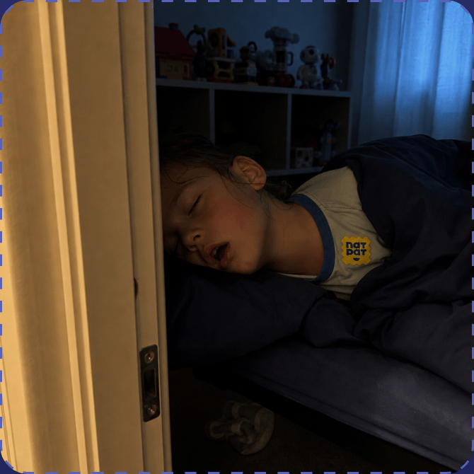 A young child asleep on their side in a dimly lit bedroom, a yellow NATPAT sticker on the shoulder of their pyjama top, seen through a partly open bedroom door.