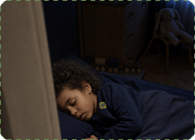 A curly-haired child asleep under a navy duvet in a dark bedroom, a small NATPAT sticker on the shoulder of their pyjama top, with a teddy bear on a chair and a chest of drawers behind.
