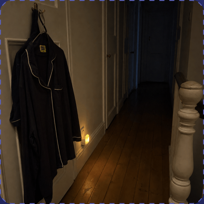 A dark upstairs hallway at night with navy pyjamas carrying a yellow NATPAT sticker hanging on a door hook, and a small nightlight glowing low on the skirting board.