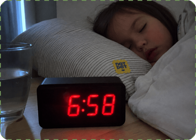 A young child asleep on a striped pillow with a yellow NATPAT sticker on it, beside a glass of water and a digital alarm clock glowing 6:58 in red.
