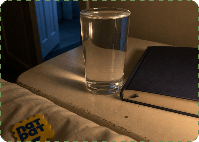 A glass of water and a closed navy notebook on a bedside table at night, a NatPat sticker on the pillow in the foreground and a lit doorway behind.