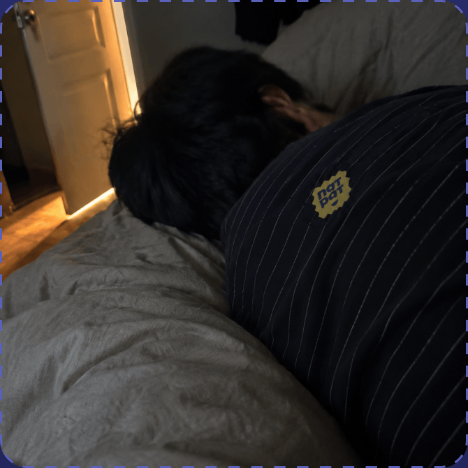 An adult asleep face-down in a dimly lit bedroom, wearing a navy pinstriped top with a yellow NATPAT sticker on the shoulder.