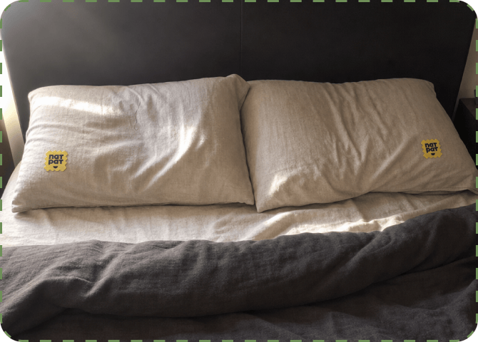 Two linen pillows side by side on a made bed, each with a yellow NATPAT sticker attached near the corner.