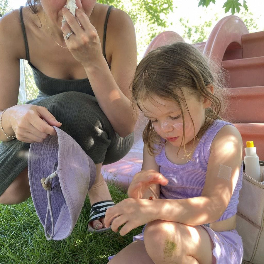 Kid proudly pointing at the patch on their own arm, parent holding sunscreen, garden light. The kid is the enforcer now.