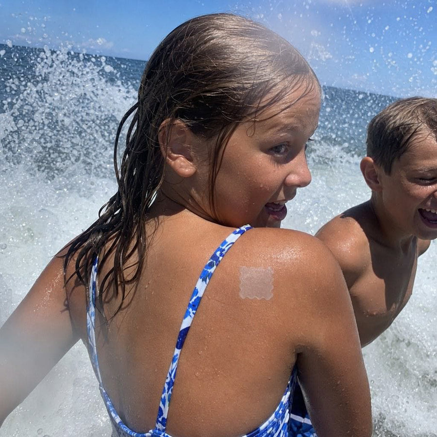Wet kid mid-laugh at the shoreline, patch visible on arm turning purple, parent with sunscreen bottle approaching.