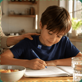 A boy in a navy t-shirt with a ZenPatch sticker near his collar writes in an exercise book at a sunlit kitchen table while a parent rests a hand on his shoulder.