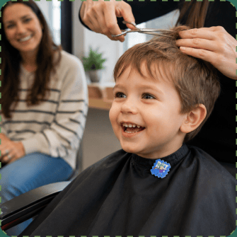 A giggling toddler in a black salon cape, a blue NatPat sticker on the cape collar, mid-haircut with the stylist's scissors above him and his smiling mother watching from behind.