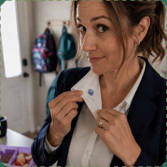A woman in a navy blazer and white shirt looks over her shoulder with a half-smile while pressing a small blue ZenPatch sticker onto her shirt collar, children's backpacks hanging on hooks by the front door behind her.